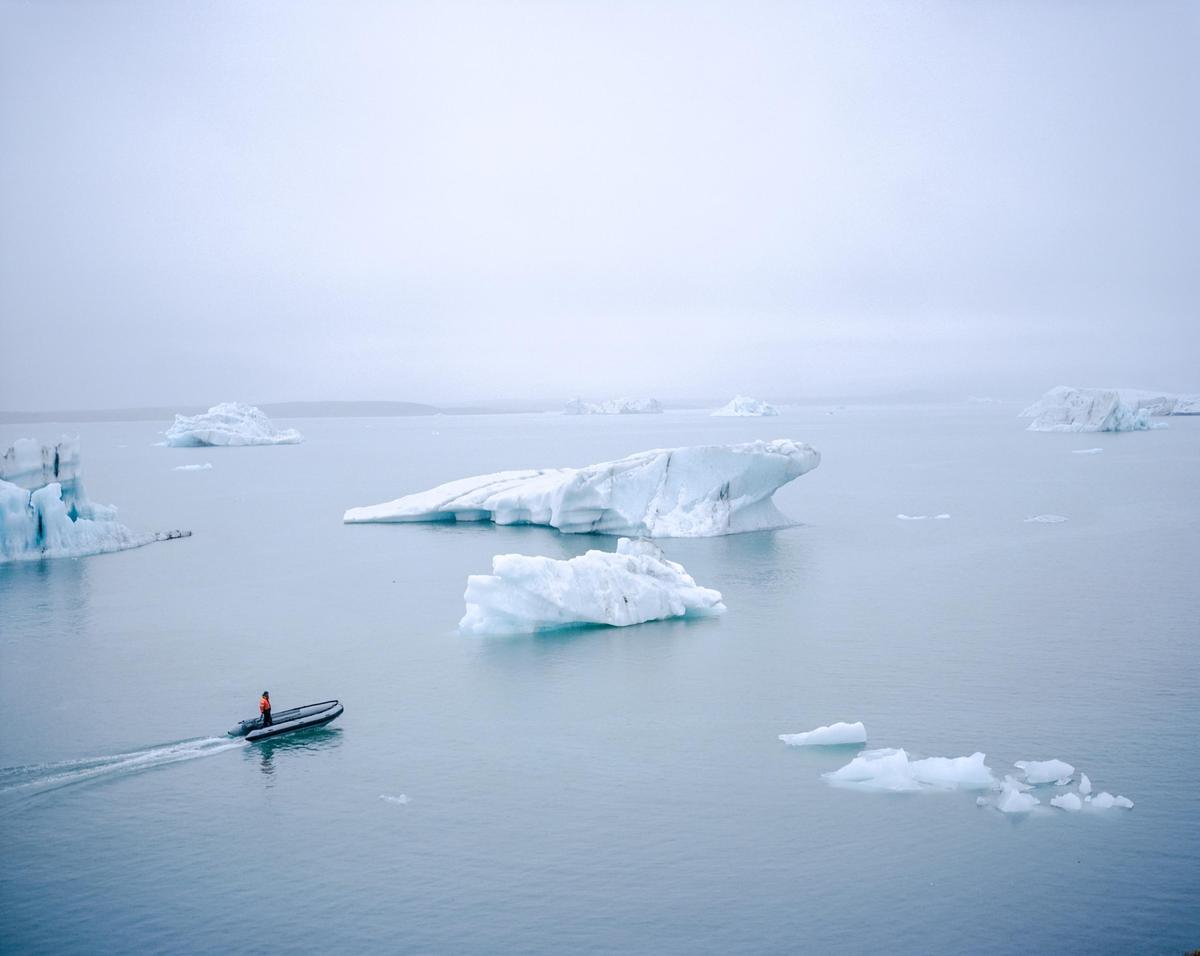 A small boat with a person in an orange jacket navigates through a body of water filled with icebergs under a hazy sky.