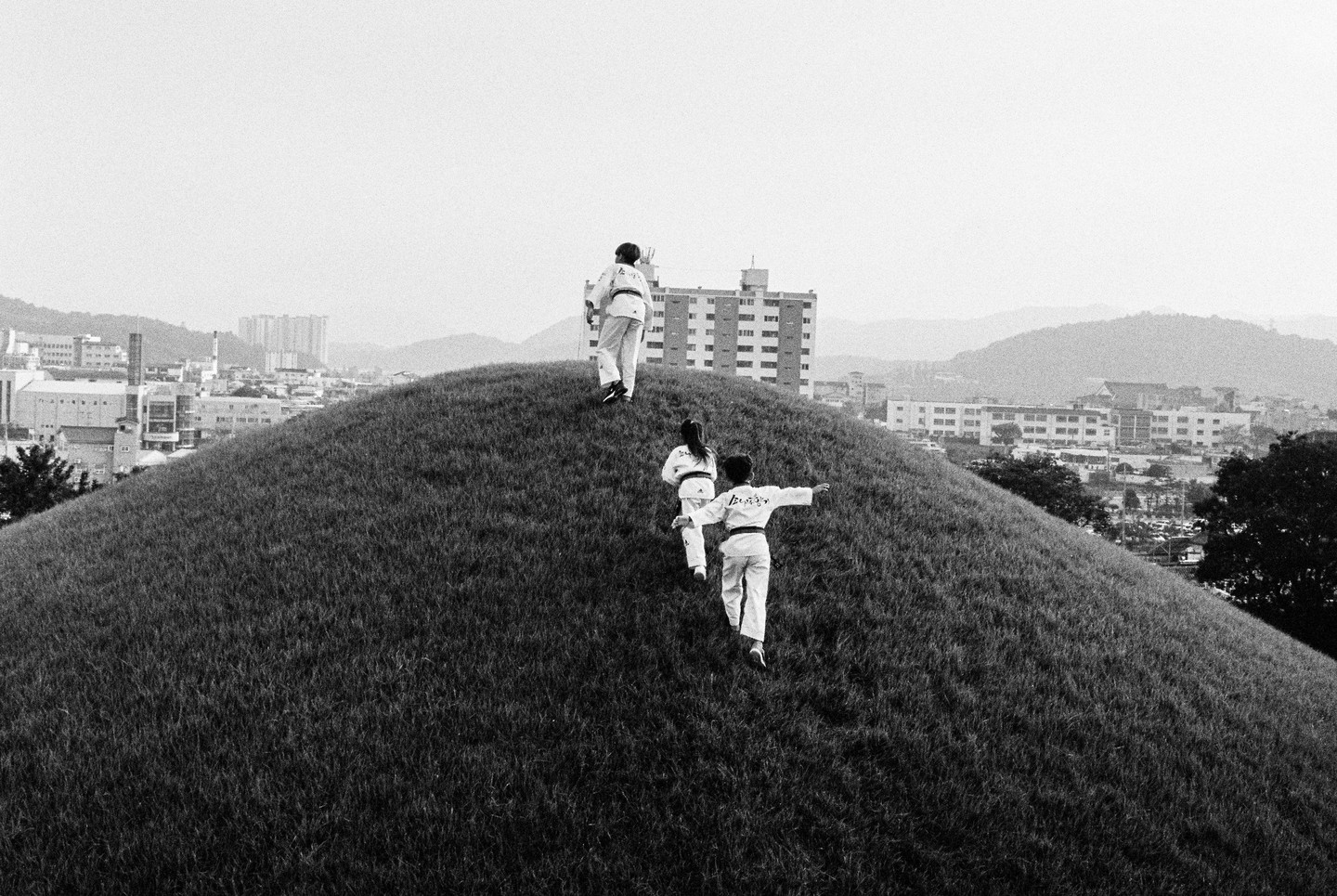 Three children in martial arts uniforms walk up a grassy hill overlooking a city.