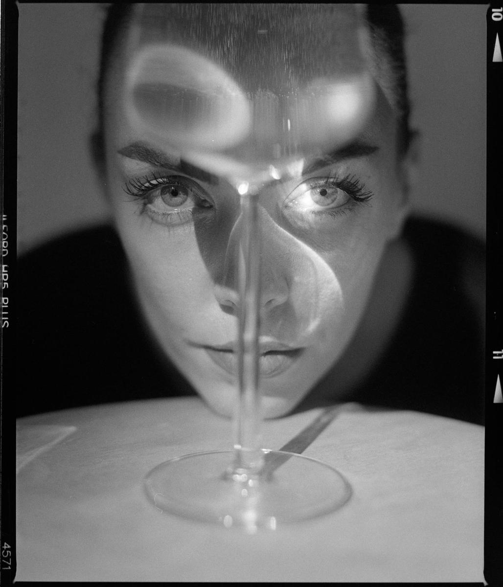 A woman's face is visible behind a wine glass, with light reflecting off the glass onto her face.