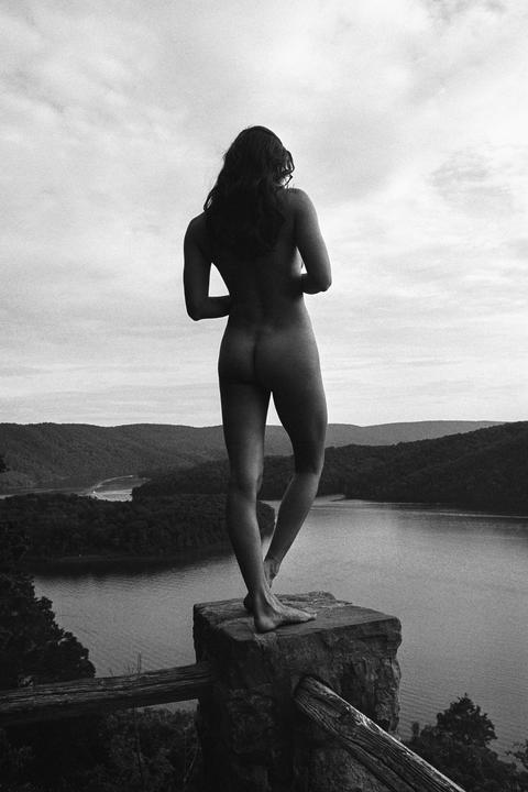 A nude woman stands on a stone pillar overlooking a lake surrounded by hills.