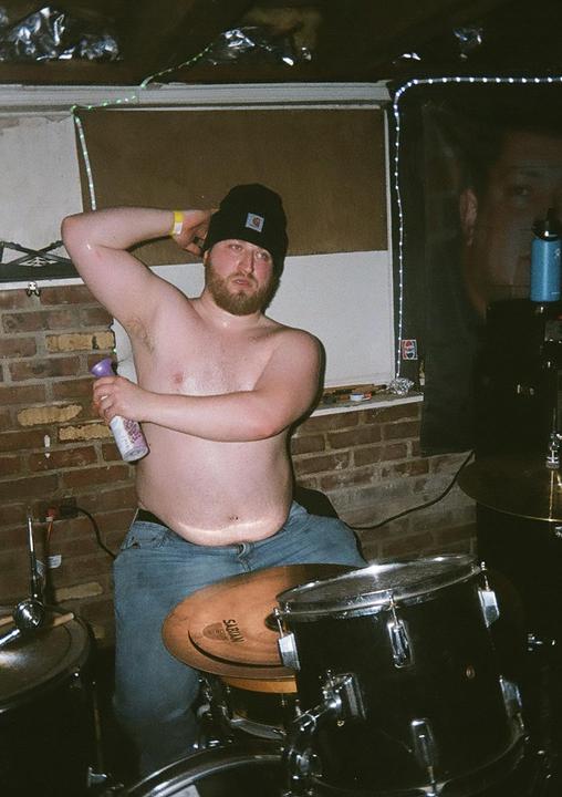 A shirtless man wearing a beanie and jeans sits at a drum kit, holding a spray bottle.