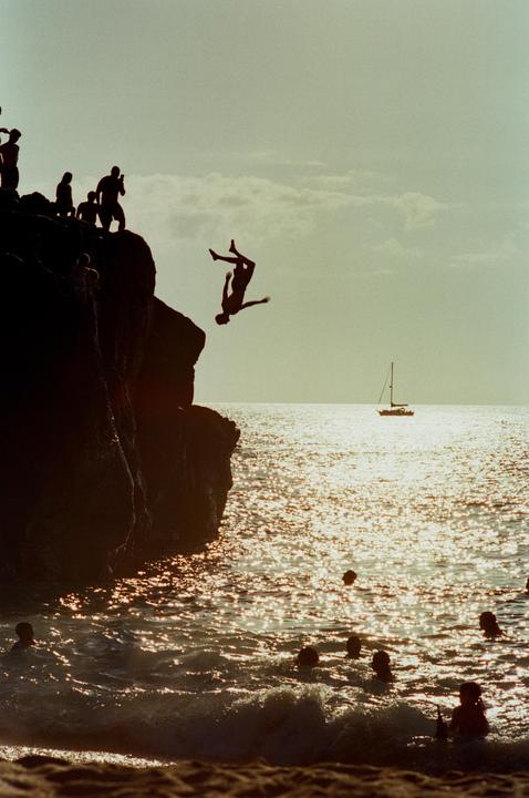 A person dives from a cliff into the ocean where other people are swimming, with a sailboat visible in the distance.