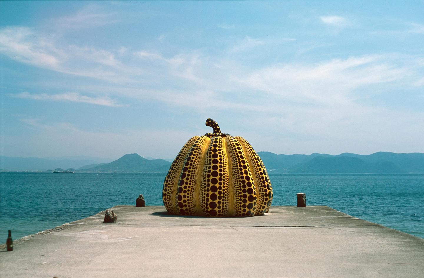A large yellow pumpkin sculpture with black polka dots sits on a concrete pier overlooking a body of water with mountains in the distance.