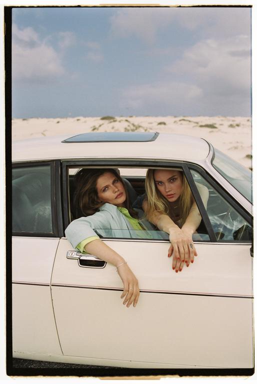 Two women are looking out of the windows of a white car in a desert landscape.