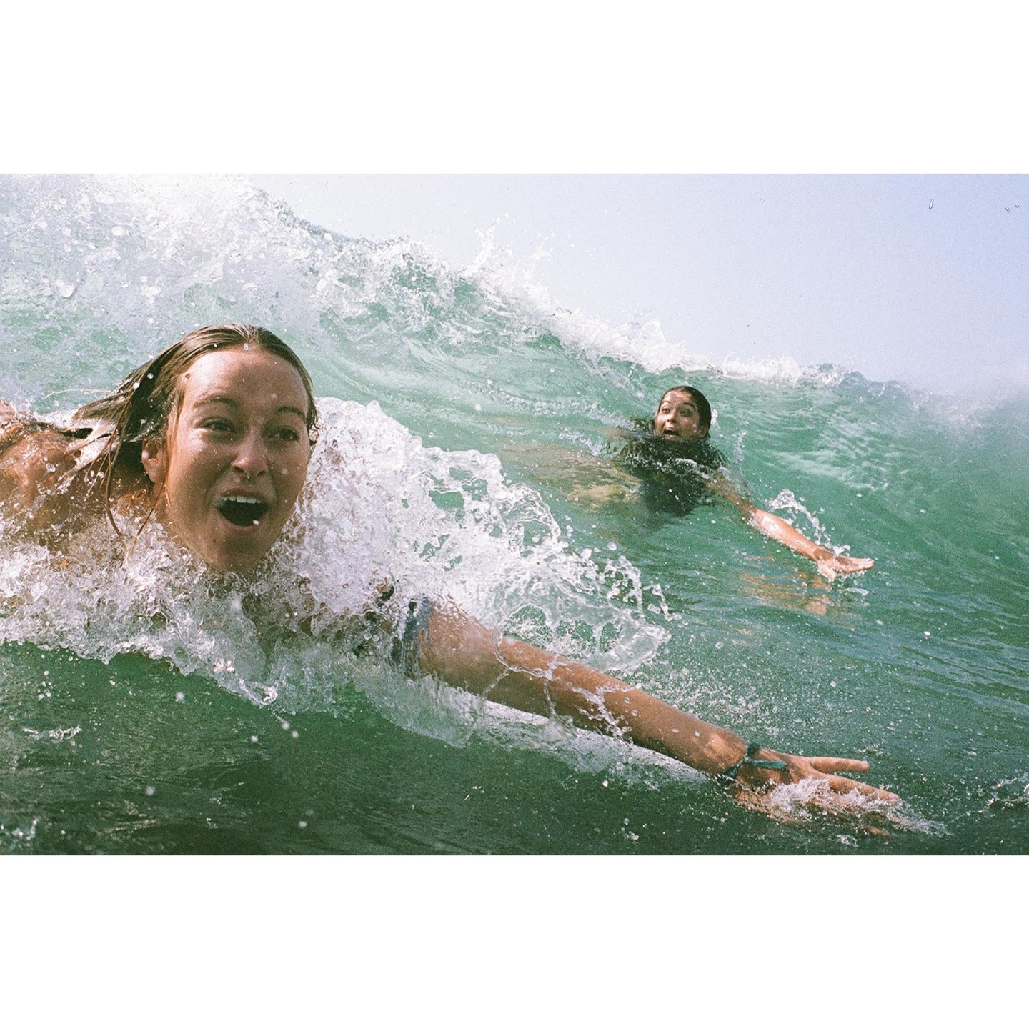 Two women are in the ocean with a large wave breaking over them.