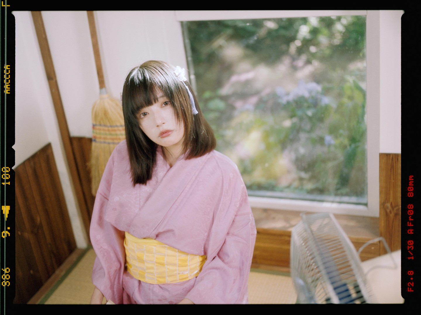 A woman in a pink kimono with a yellow obi sits on a tatami mat next to a window, with a fan and brooms visible.