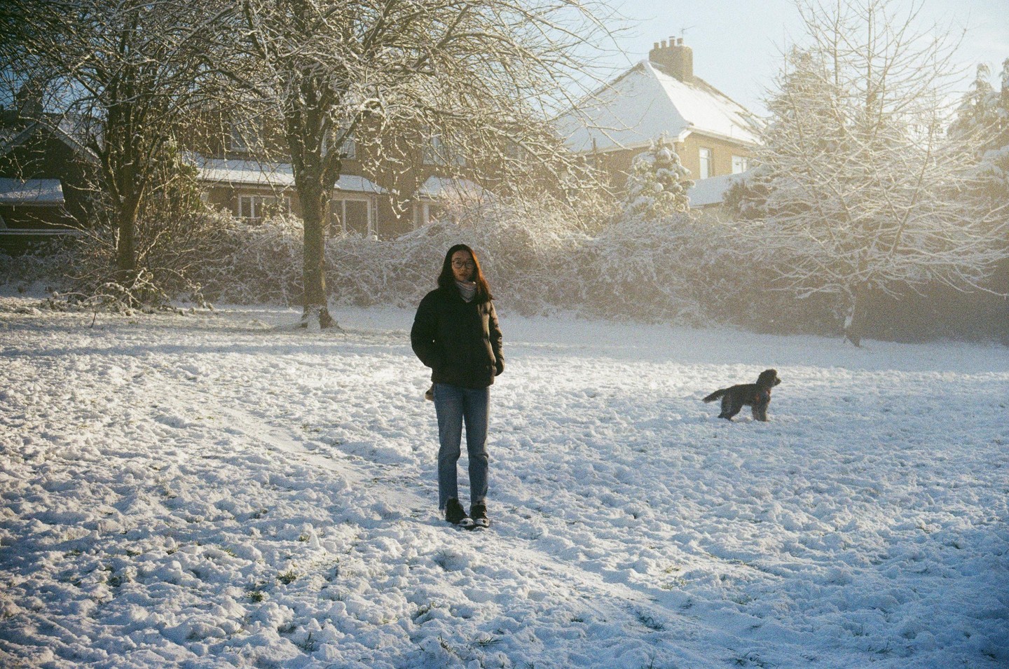 A woman stands in a snowy field with a dog and houses in the background.