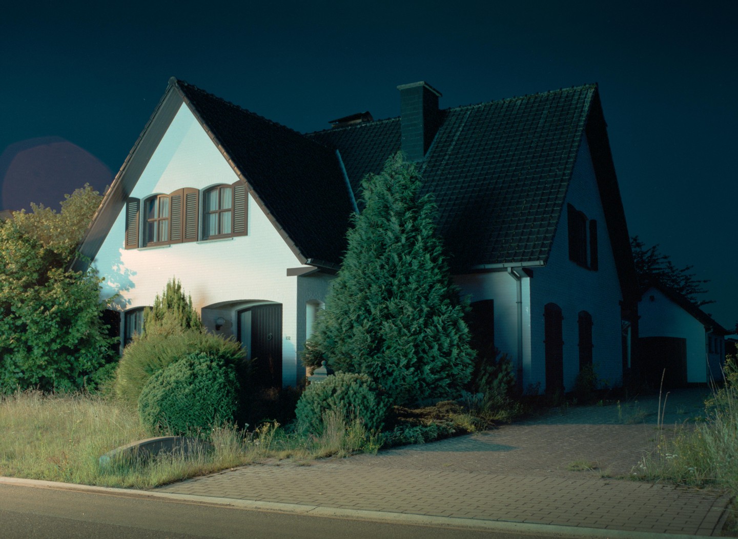 A white house with a dark roof and brown shutters is illuminated at night, surrounded by trees and bushes.