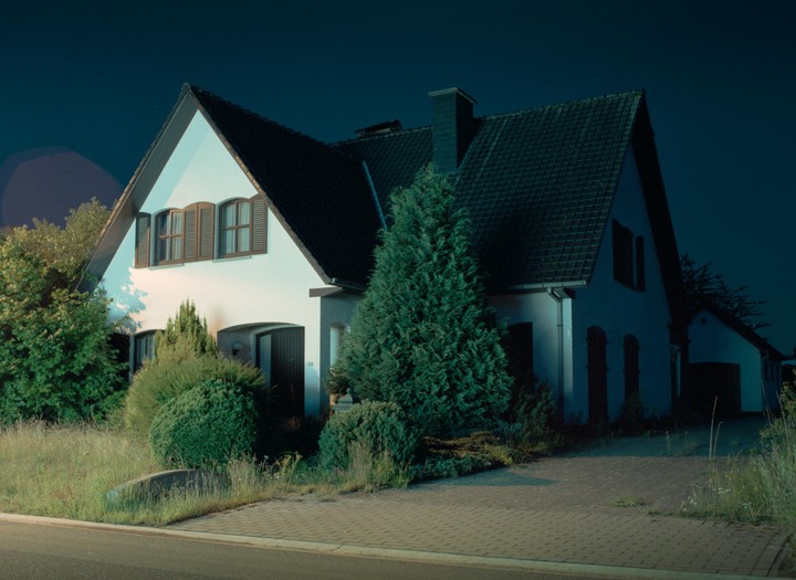 A white house with a dark roof and brown shutters is illuminated at night, surrounded by trees and bushes.