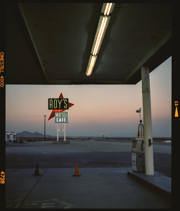 A neon sign for Roy's Motel and Cafe stands in a desert landscape at dusk, with a gas pump and traffic cones in the foreground.
