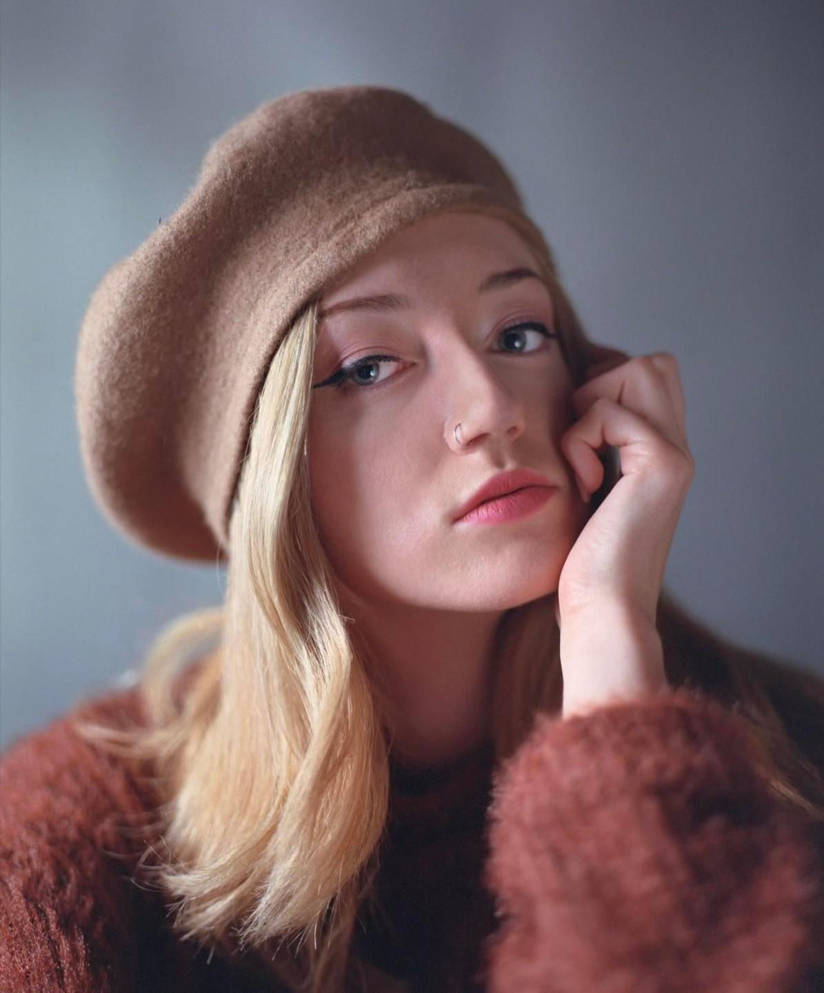 A woman wearing a beret and a fuzzy sweater poses for a portrait with her hand resting on her chin.