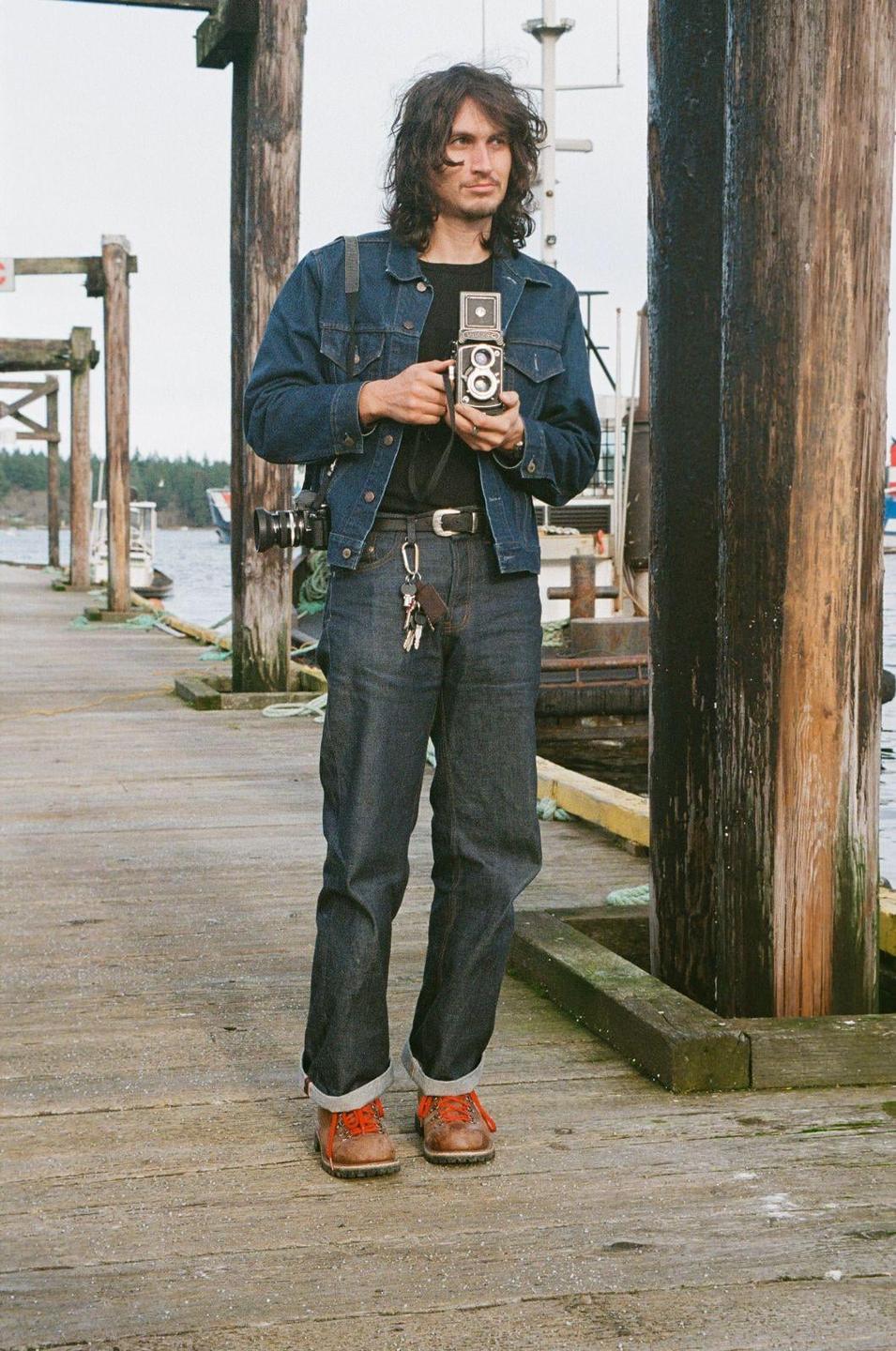 A man stands on a wooden pier holding a camera, with water and boats in the background.