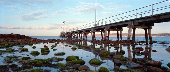A long pier extends over a rocky, moss-covered shoreline with calm water reflecting the sky.