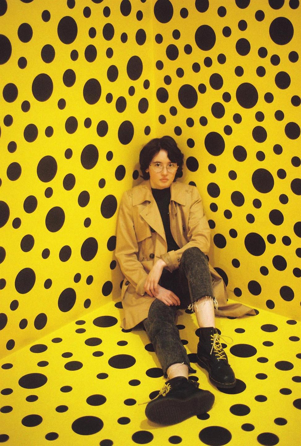 A woman wearing a trench coat and glasses sits in a yellow room with black polka dots.