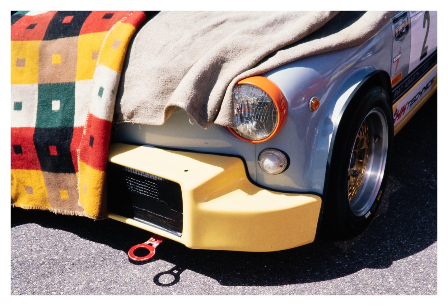 A light blue car with a yellow bumper and orange headlight trim is partially covered by a colorful blanket and a beige cloth.