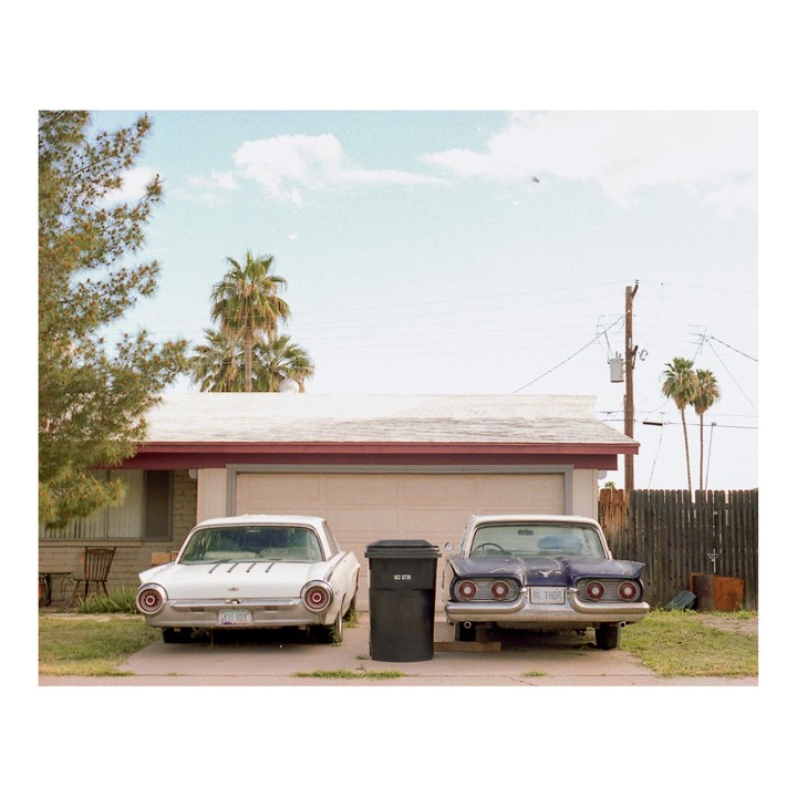 Two vintage cars are parked in front of a garage with palm trees in the background.