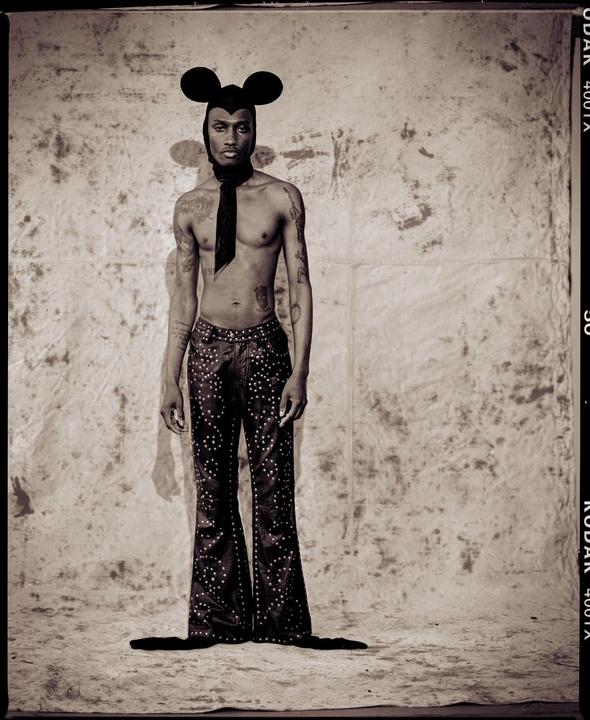A man wearing a Mickey Mouse hat, a scarf, and studded bell-bottom pants stands in front of a textured backdrop.