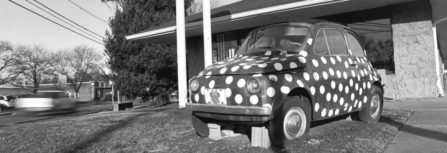 A black and white photograph shows a small car with white polka dots parked on a grassy area next to a building, with a blurred car passing on the street in the background.