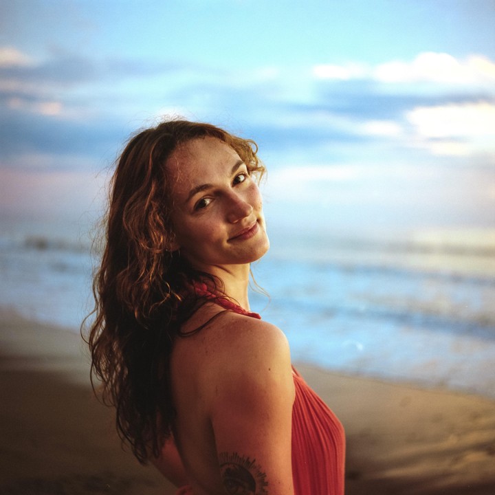 A woman with long, curly hair looks over her shoulder at the camera while standing on a beach at dusk.