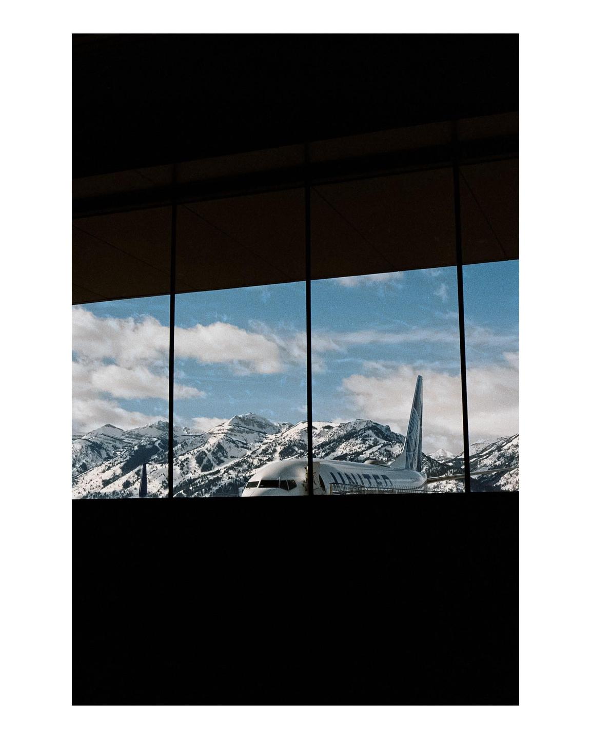 An airplane is visible through a window with snow-capped mountains in the background.