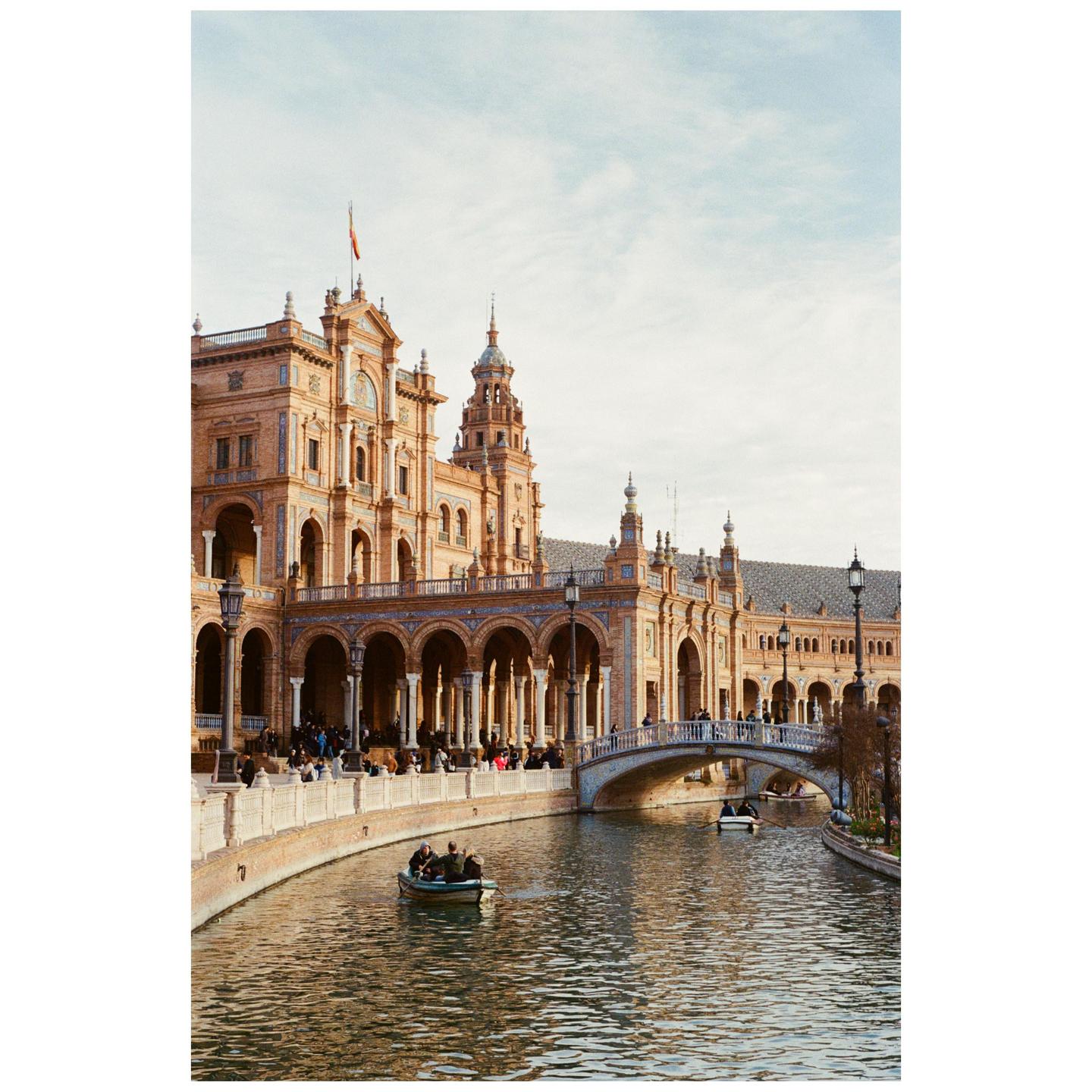 A large, ornate building with arches and towers stands beside a canal with people in boats and on a bridge.