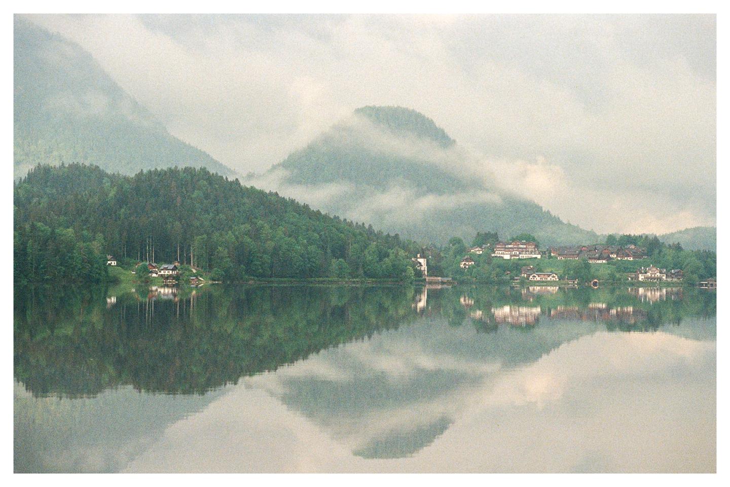 A village with houses and trees is reflected in a lake with mountains in the background.