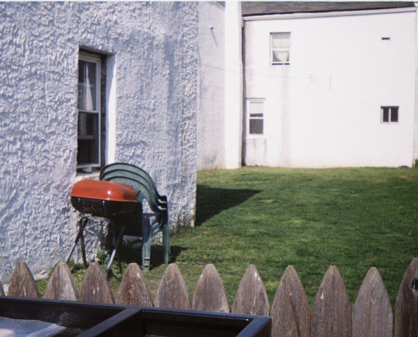 A red grill and stacked green chairs sit beside a white stucco building with a wooden fence in the foreground.