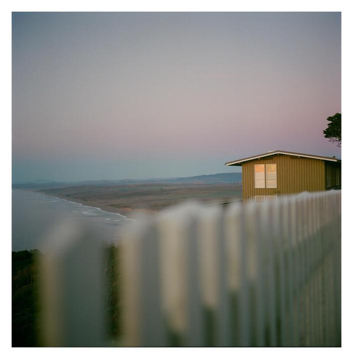 A house sits on a cliff overlooking a beach and ocean, with a white picket fence in the foreground.