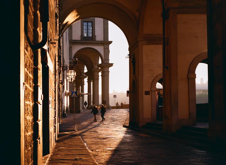 Two people walk on a sunlit cobblestone street under an archway in Florence.
