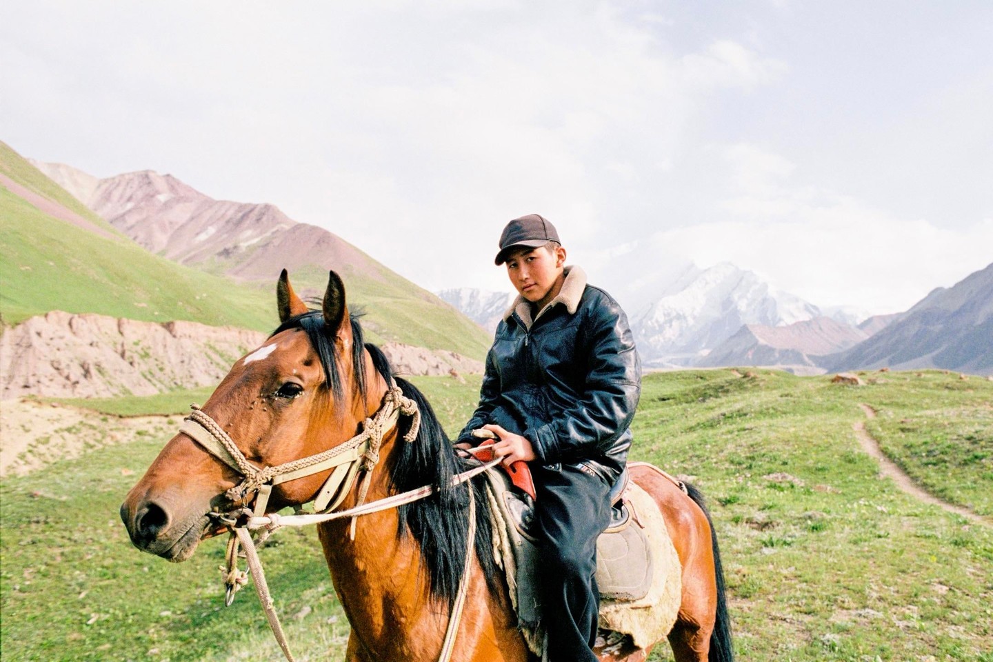 A boy on horseback looks at the viewer with mountains and green hills in the background.