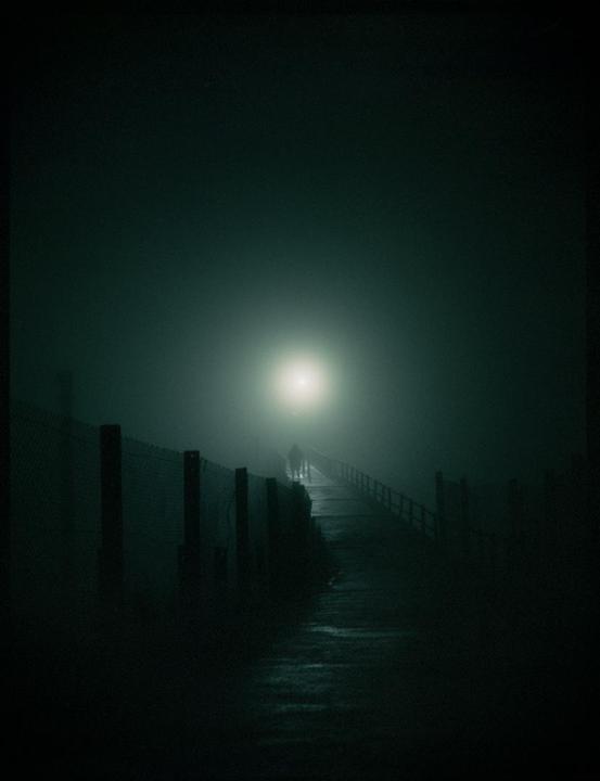 Two people walk on a foggy path at night, illuminated by a bright light in the distance.