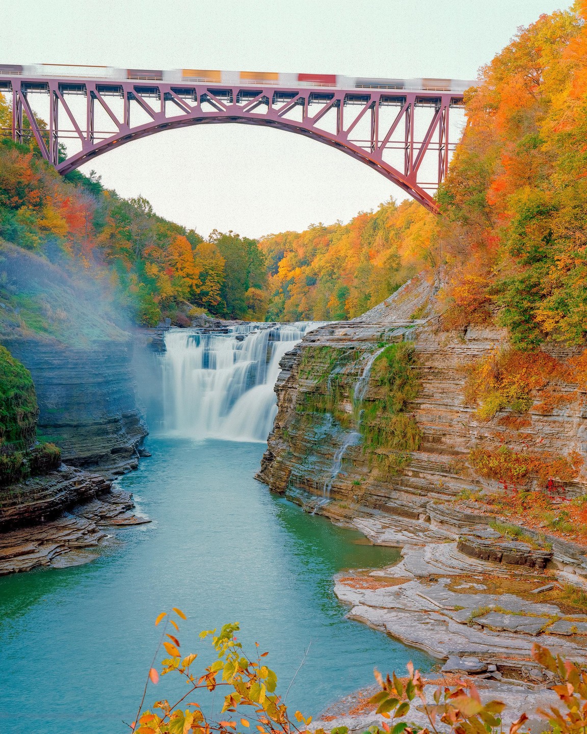 A train crosses a bridge over a river with a waterfall and autumn trees.