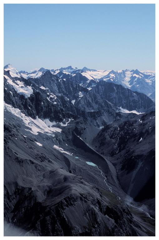 A vast mountain range with snow-capped peaks and a valley with a moraine and a small turquoise lake is visible under a clear sky.
