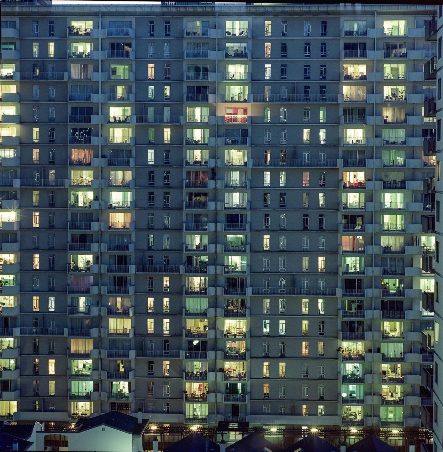 A tall apartment building with many windows lit up at night stands above a row of houses.