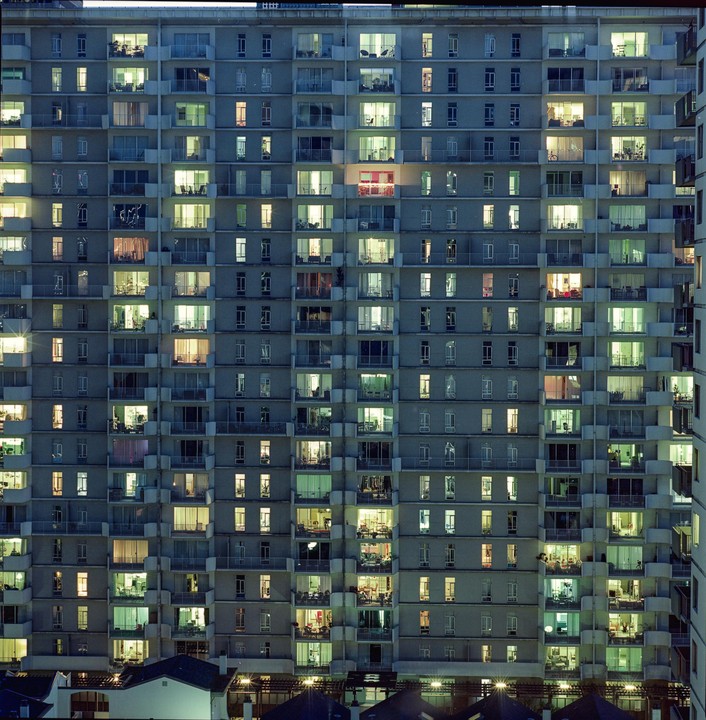 A tall apartment building with many windows lit up at night stands above a row of houses.