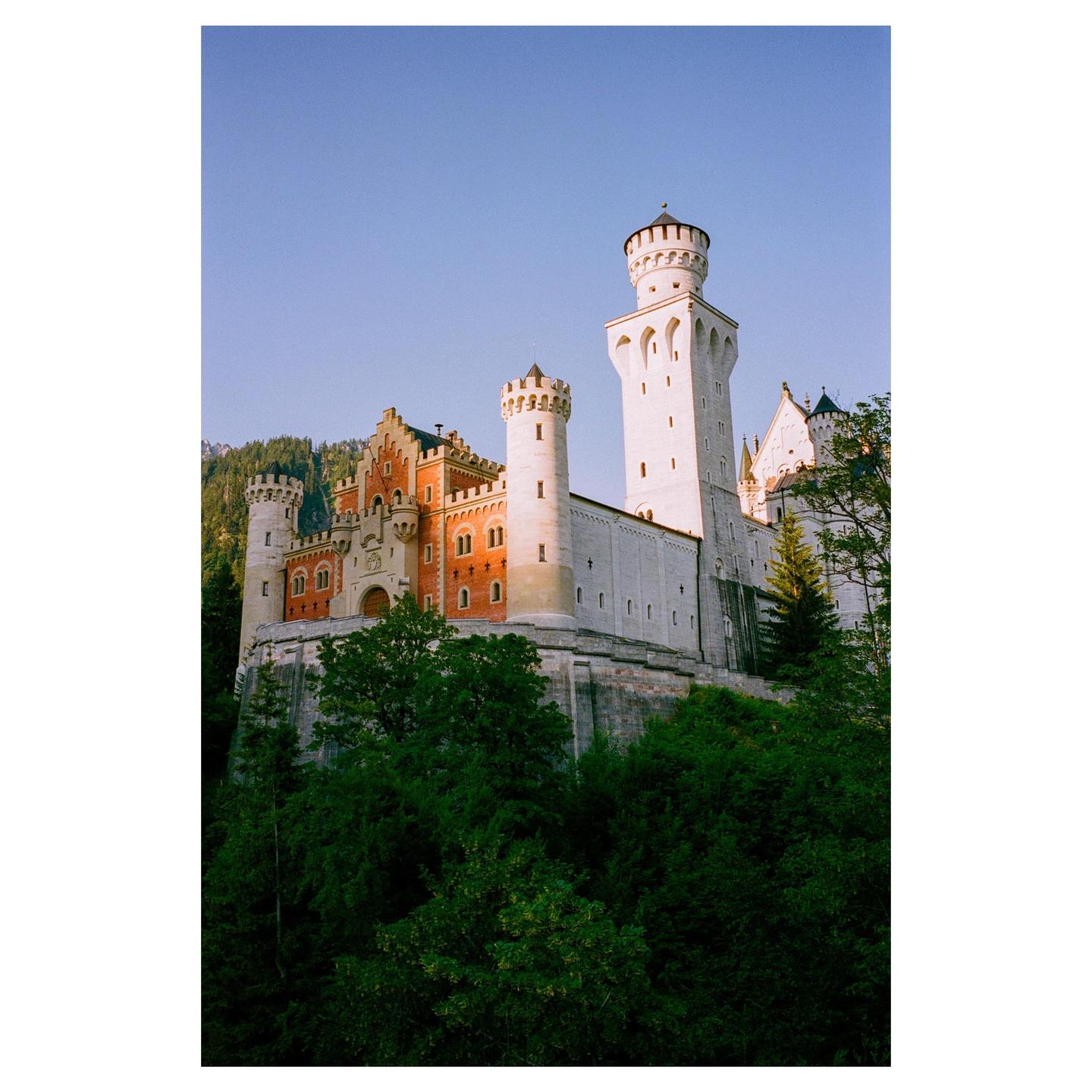 A large castle with multiple towers and red roofs stands on a hill surrounded by green trees under a clear blue sky.