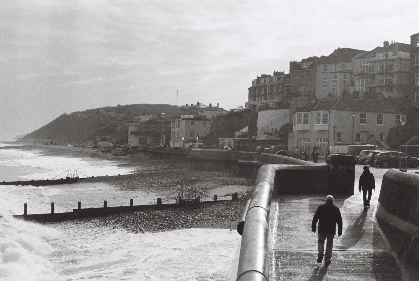 Two men walk along a concrete promenade next to a beach with waves crashing against groynes, with a town built into a hillside in the background.