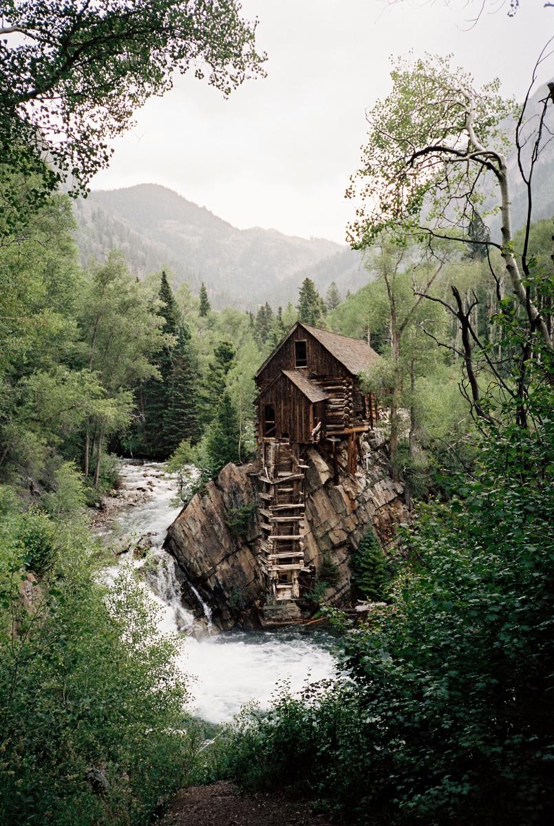 A wooden mill sits on a rocky outcrop next to a river with a waterfall, surrounded by a forest and mountains.