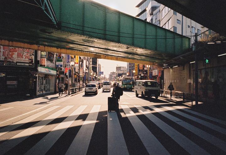 A person pulls a suitcase across a crosswalk under a green overpass in a city street with cars and buildings.