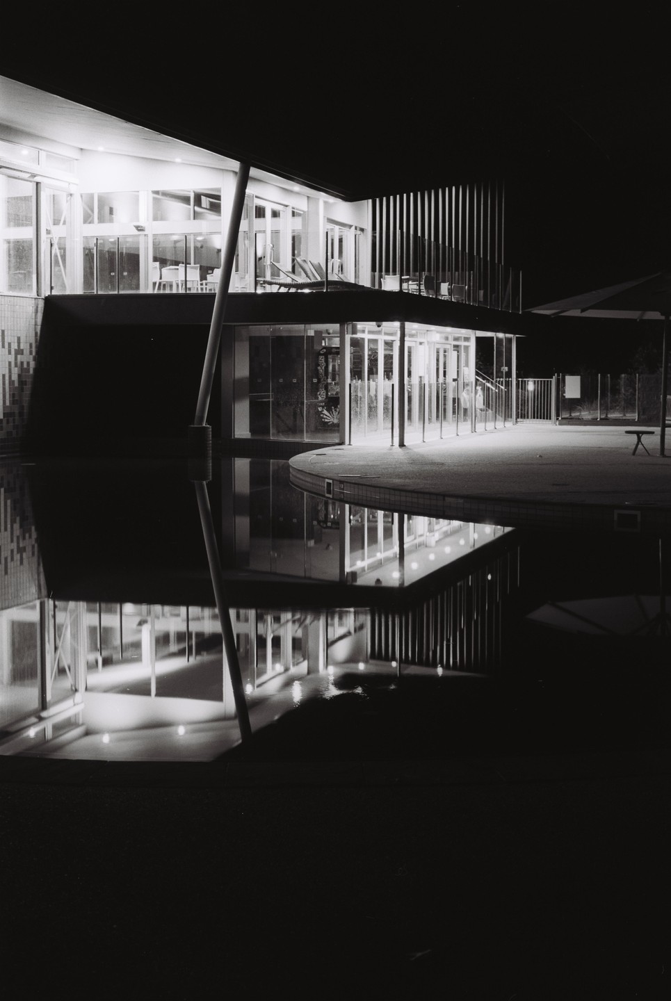 A modern building with large windows is illuminated at night, reflecting in the still water of a pool or pond in the foreground.