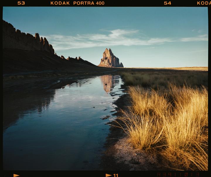 A large rock formation is reflected in a body of water next to a field of dry grass.
