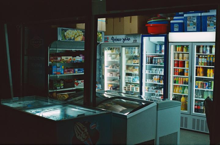 A dark supermarket interior is illuminated by the bright lights of several refrigerators and freezers.