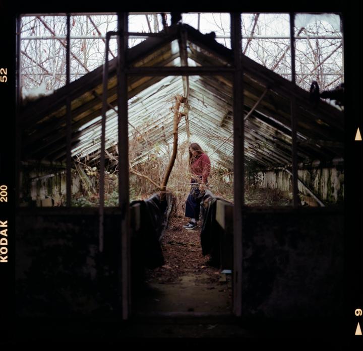 A woman stands inside a dilapidated greenhouse with broken glass panels and overgrown plants.