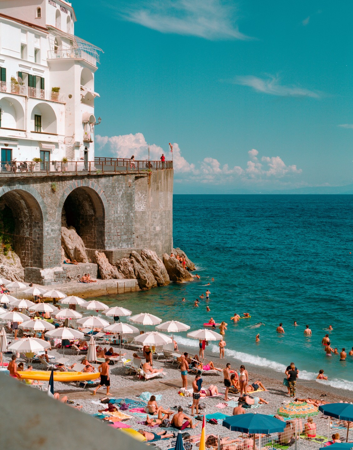 A white hotel building with arched foundations overlooks a crowded beach and the sea.