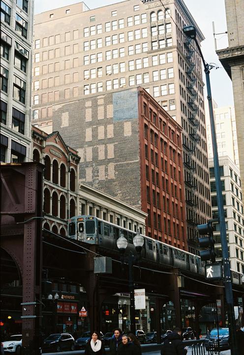 A train travels on an elevated track through a city street lined with tall buildings.