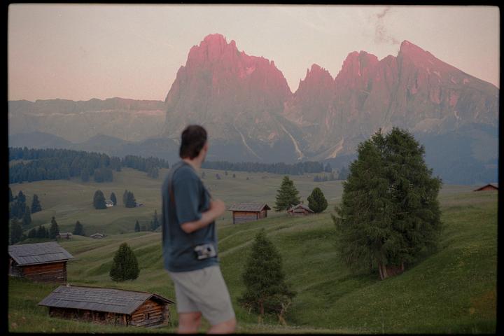 A man stands in a grassy field with wooden huts and evergreen trees, looking at pink-hued mountains in the distance.