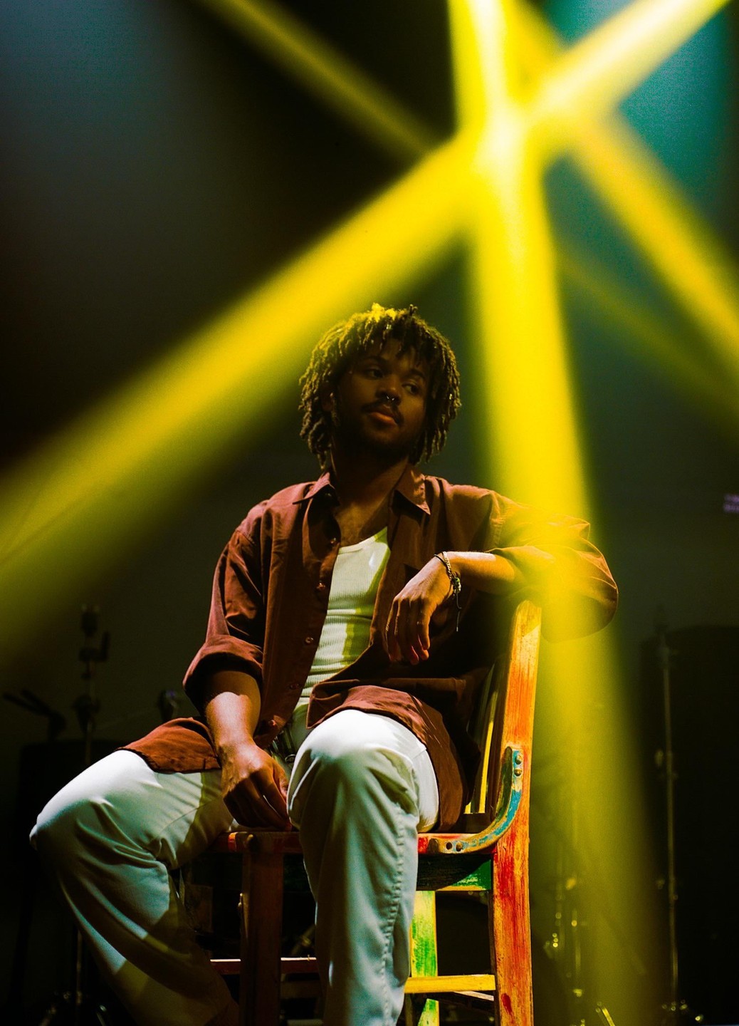 A man with dreadlocks and a nose ring sits on a colorful chair under bright yellow stage lights.