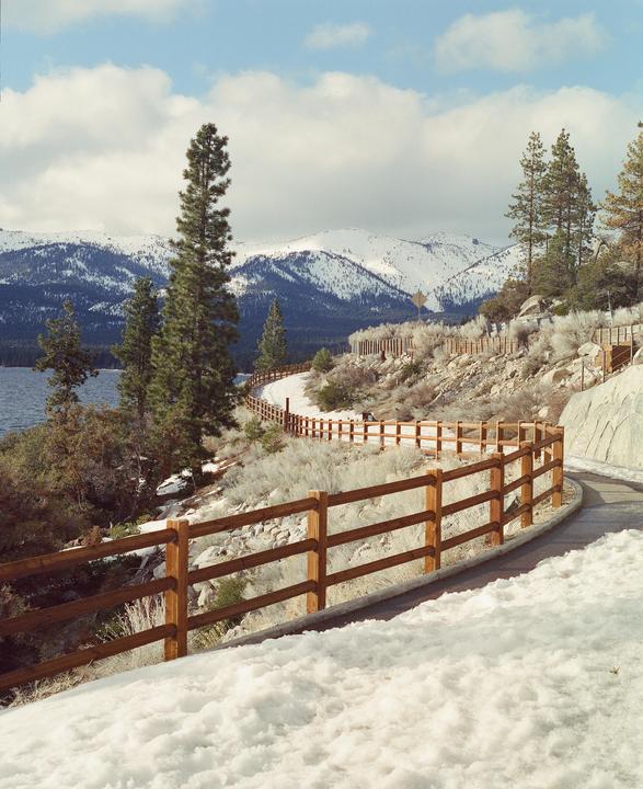A winding path with a wooden fence runs alongside a lake, with snow-capped mountains and pine trees in the background.
