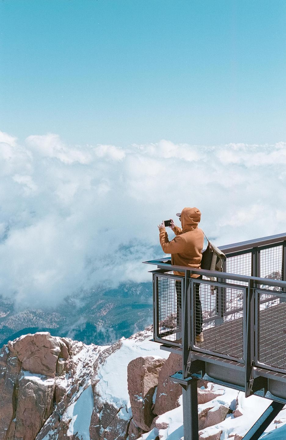 A person stands on a metal observation deck, taking a photo of a vast cloudscape and snow-covered mountains.