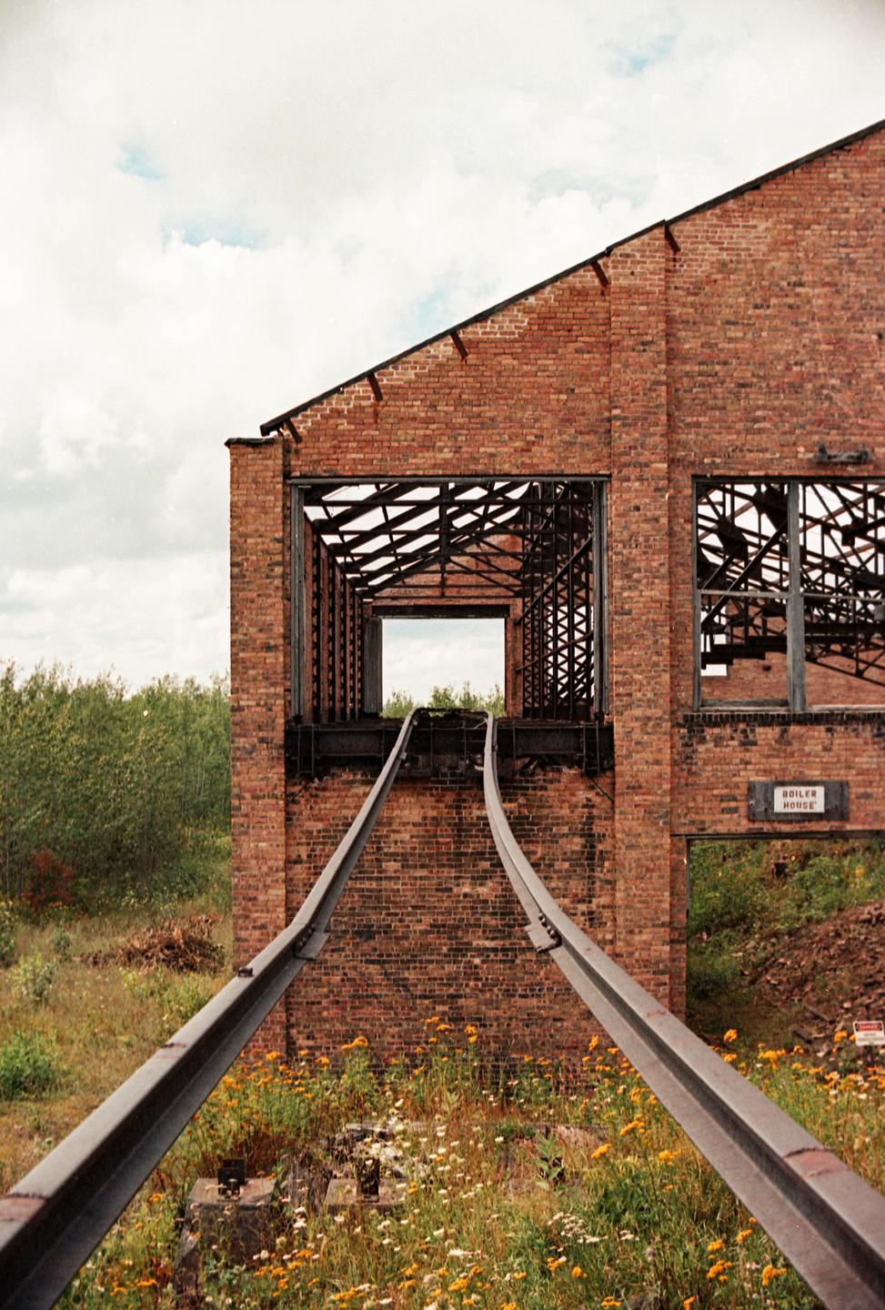 A brick boiler house with a collapsed roof and two metal tracks leading into it is surrounded by wildflowers and trees under a cloudy sky.